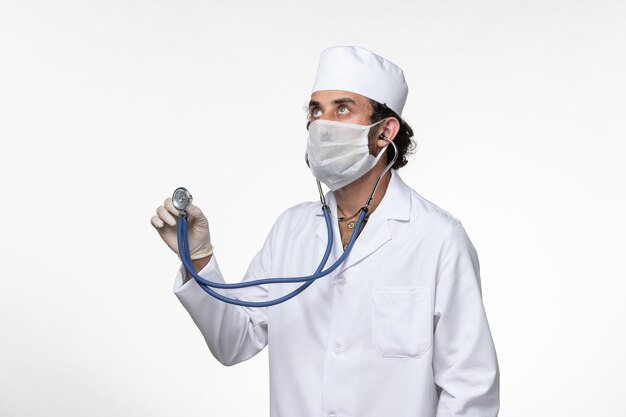 Front view male doctor in medical suit and wearing a mask as a protection from covid- using stethoscope on the white wall illness virus covid- pandemic