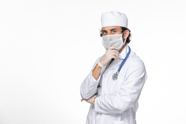 Front view male doctor in medical suit wearing sterile mask as a protection from covid posing and thinking on white wall splash virus coronavirus pandemic health