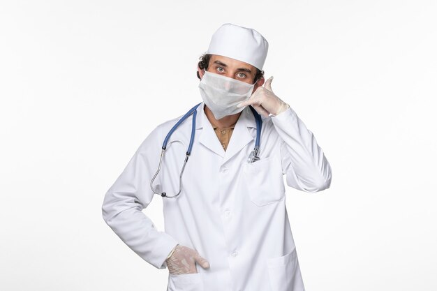 Front view male doctor in medical suit wearing sterile mask as a protection from covid showing phone call pose on white desk virus coronavirus pandemic disease