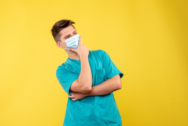 Front view of male doctor in medical suit with sterile mask thinking on yellow wall