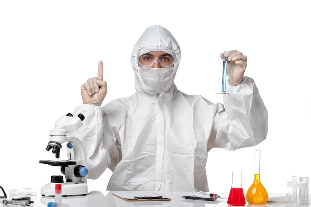 Front view male doctor in protective suit with mask due to covid holding flask with blue solution on a white space