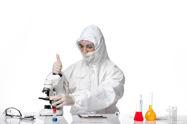 Front view male doctor in protective suit with mask due to covid using microscope on the light-white space