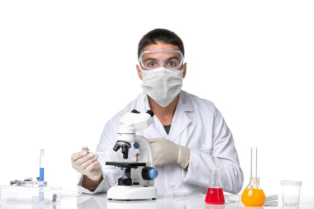 Front view male doctor in white medical suit with mask due to covid using microscope on light-white space