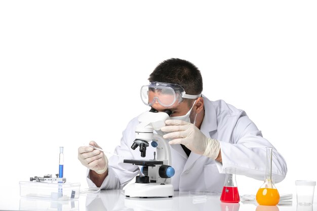 Front view male doctor in white medical suit with mask due to covid using microscope on a white space