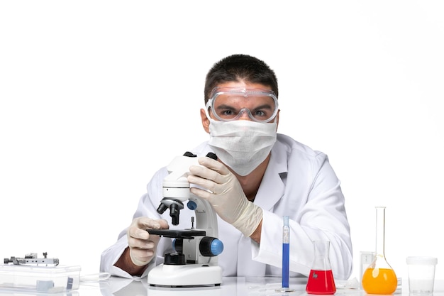 Front view male doctor in white medical suit with mask due to covid using microscope on the white space