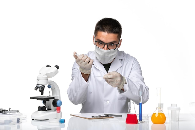 Front view male doctor in white medical suit with mask due to covid working with solutions on white desk