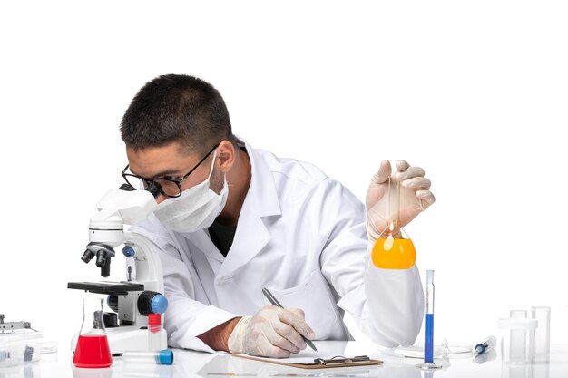 Front view male doctor in white medical suit and with mask working with solutions on a white space