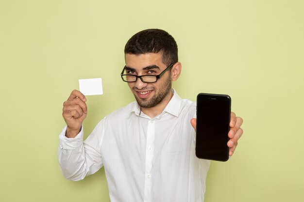 Front view of male office worker in white shirt holding smartphone and card on light-green wall