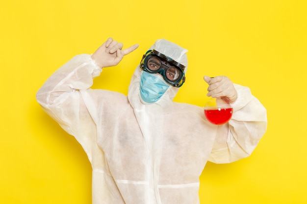 Front view male scientific worker in special protective suit holding flask with red solution posing on light yellow desk