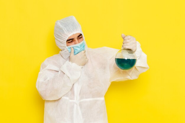 Front view of male scientific worker in special protective suit with mask holding flask thinking on yellow wall