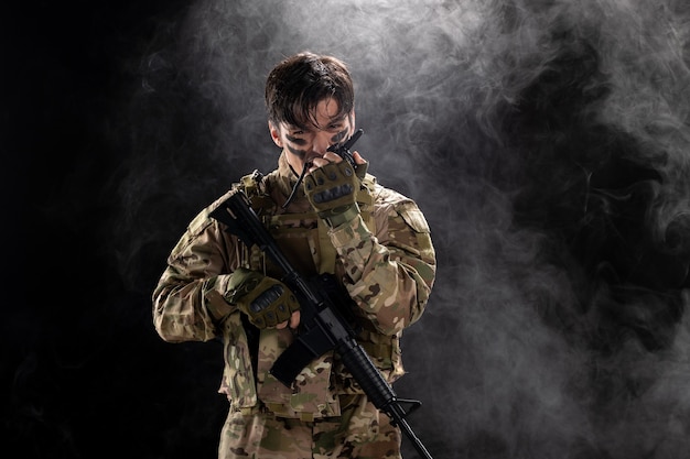 Front view of male soldier with machine gun using walkie-talkie black wall