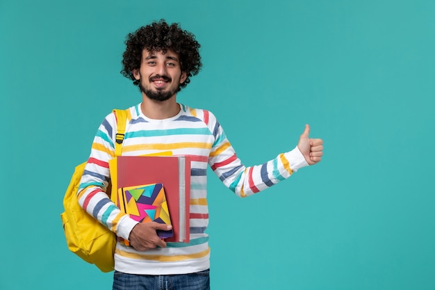 Front view of male student in colored striped shirt wearing yellow backpack holding files and copybooks on blue wall