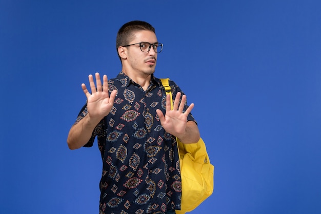 Front view of male student in dark cotton shirt wearing yellow backpack on blue wall