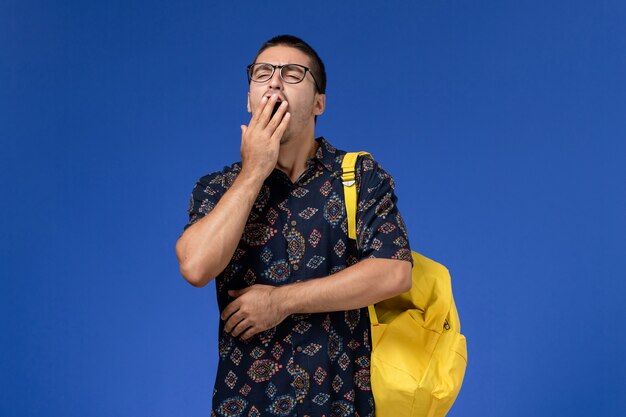 Front view of male student in dark shirt wearing yellow backpack yawning on the blue wall