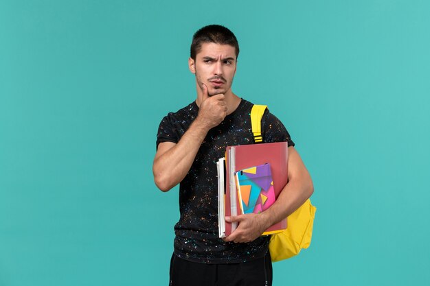 Front view of male student in dark t-shirt wearing yellow backpack holding copybook and files thinking on blue wall