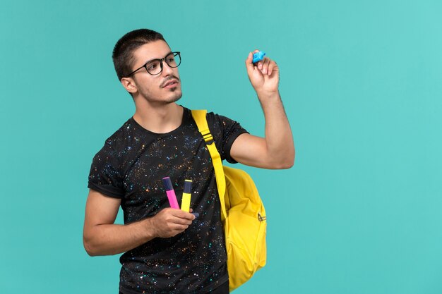 Front view of male student in dark t-shirt yellow backpack holding colored felt pens on blue wall