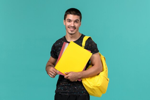 Front view of male student in dark t-shirt yellow backpack holding different files on light blue wall