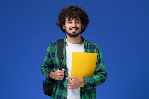 Front view of male student in green checkered shirt wearing black backpack and holding files on blue wall