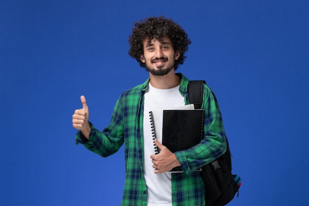 Front view of male student in green checkered shirt with black backpack holding copybooks smiling on blue wall