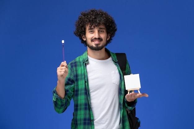 Free photo front view of male student in green checkered shirt with black backpack holding tassel and easel on blue wall