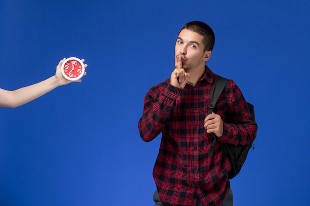 Front view of male student in red checkered shirt with backpack on blue wall