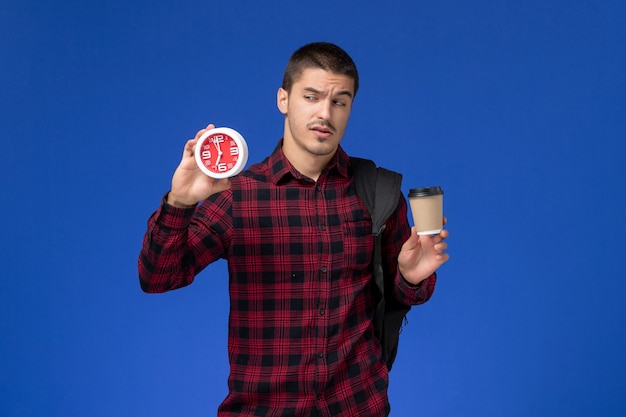 Front view of male student in red checkered shirt with backpack holding clocks and coffee on the blue wall