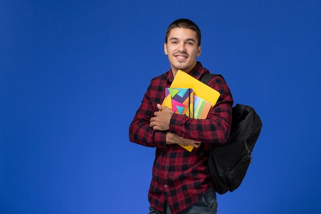 Front view of male student in red checkered shirt with backpack holding copybook and files on the light-blue wall