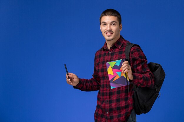 Front view of male student in red checkered shirt with backpack holding copybook and pen on light-blue wall