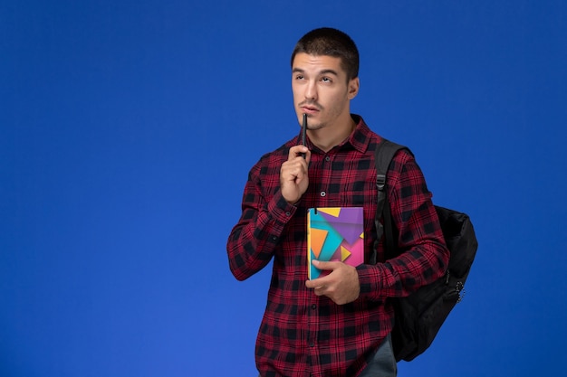 Front view of male student in red checkered shirt with backpack holding copybook and thinking on light-blue wall