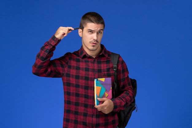 Front view of male student in red checkered shirt with backpack holding copybook thinking on the light blue wall