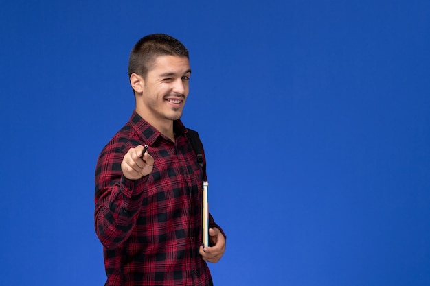 Front view of male student in red checkered shirt with backpack holding copybook winking on light-blue wall