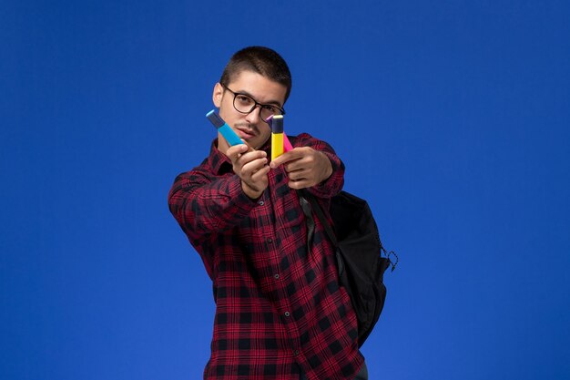 Front view of male student in red checkered shirt with backpack holding felt pens on light-blue wall