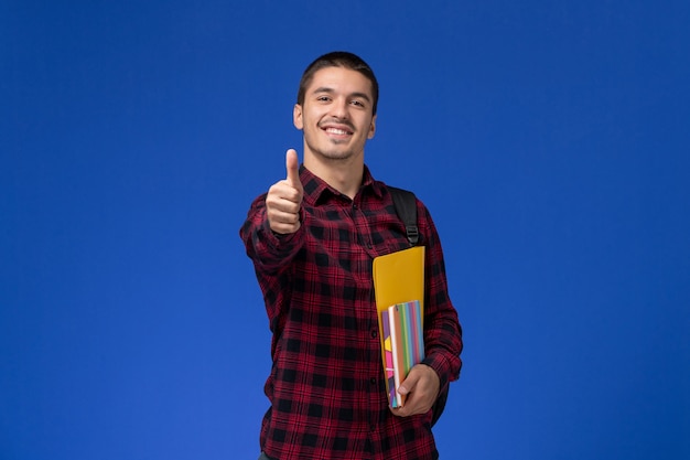 Front view of male student in red checkered shirt with backpack holding files and copybooks smiling on blue wall