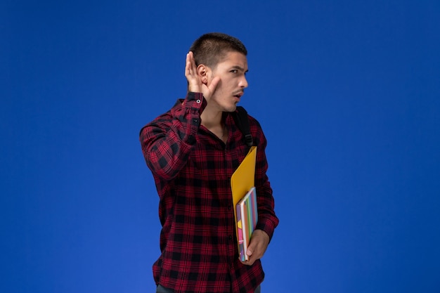 Front view of male student in red checkered shirt with backpack holding files and copybooks trying to hear on blue wall