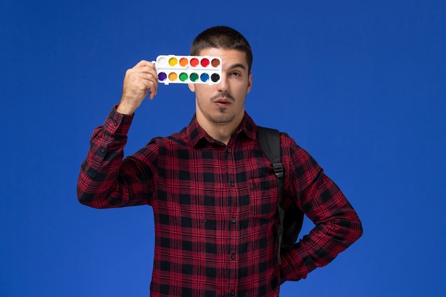 Free photo front view of male student in red checkered shirt with backpack holding paints for drawing on blue wall