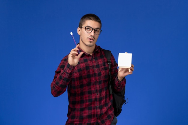 Free Photo front view of male student in red checkered shirt with backpack holding tassel and easel on blue wall