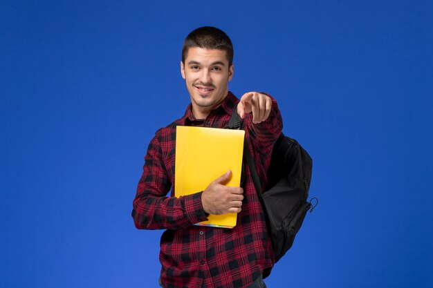 Front view of male student in red checkered shirt with backpack holding yellow files on the blue wall