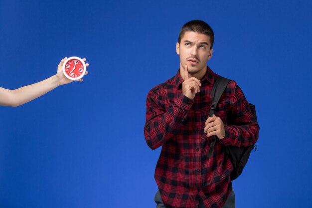 Front view of male student in red checkered shirt with backpack posing and thinking on the blue wall