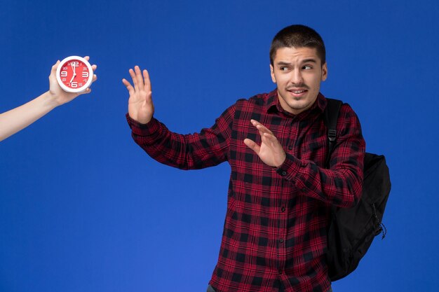 Front view of male student in red checkered shirt with backpack scared of clocks on blue wall