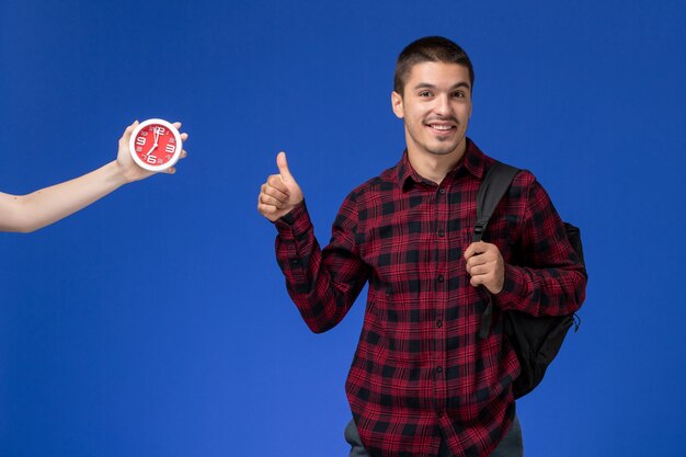 Front view of male student in red checkered shirt with backpack smiling on blue wall