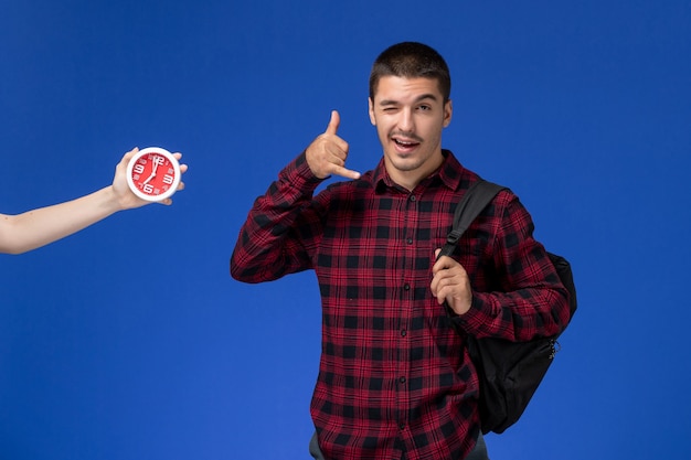 Front view of male student in red checkered shirt with backpack winking on blue wall