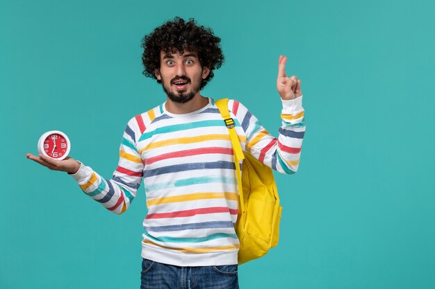 Front view of male student in striped shirt wearing yellow backpack holding clocks crossing his fingers on blue wall