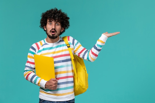 Front view of male student in striped shirt wearing yellow backpack holding files on blue wall