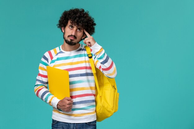 Front view of male student in striped shirt wearing yellow backpack holding files thinking on blue wall