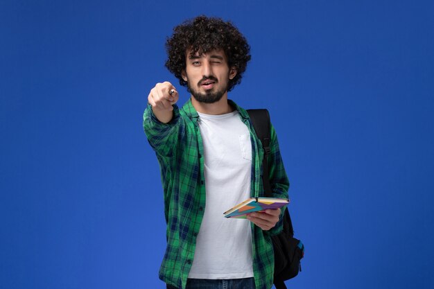 Front view of male student wearing black backpack holding copybook and pen on blue wall