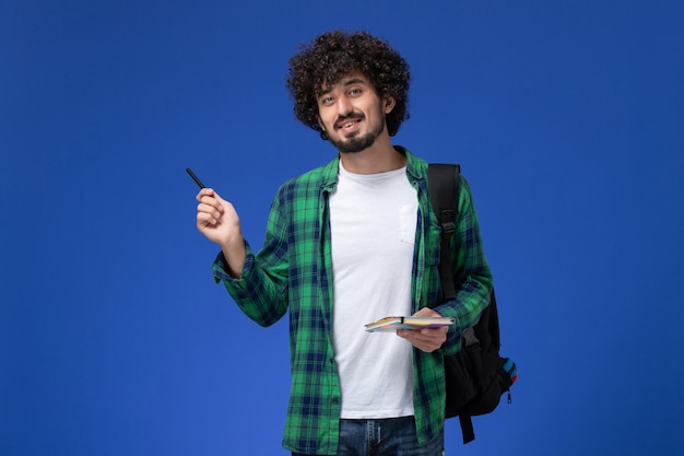 Front view of male student wearing black backpack holding copybook and pen on the blue wall