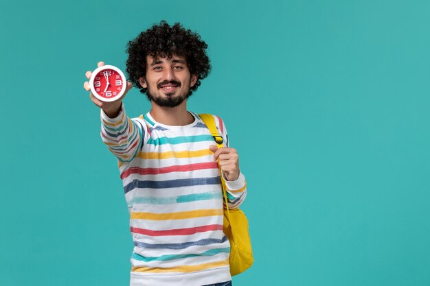 Front view of male student wearing yellow backpack holding clocks on light blue wall