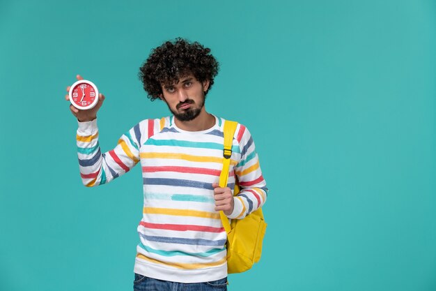 Front view of male student wearing yellow backpack holding clocks on the light blue wall