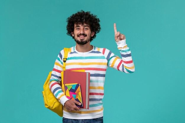 Front view of male student wearing yellow backpack holding files and copybook on blue wall