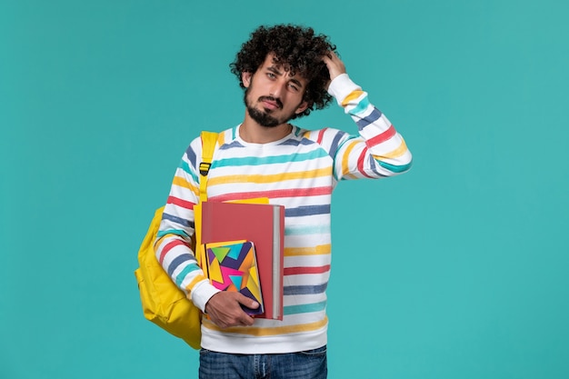 Front view of male student wearing yellow backpack holding files and copybook on blue wall